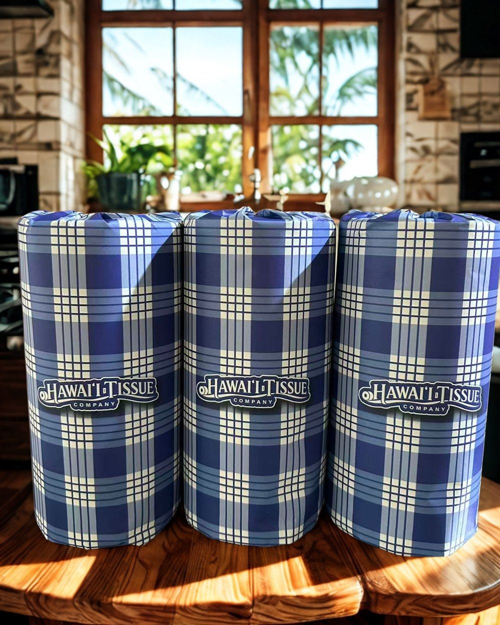 Three palaka-wrapped bamboo paper towel rolls on a wood table in a bright Hawaiian kitchen