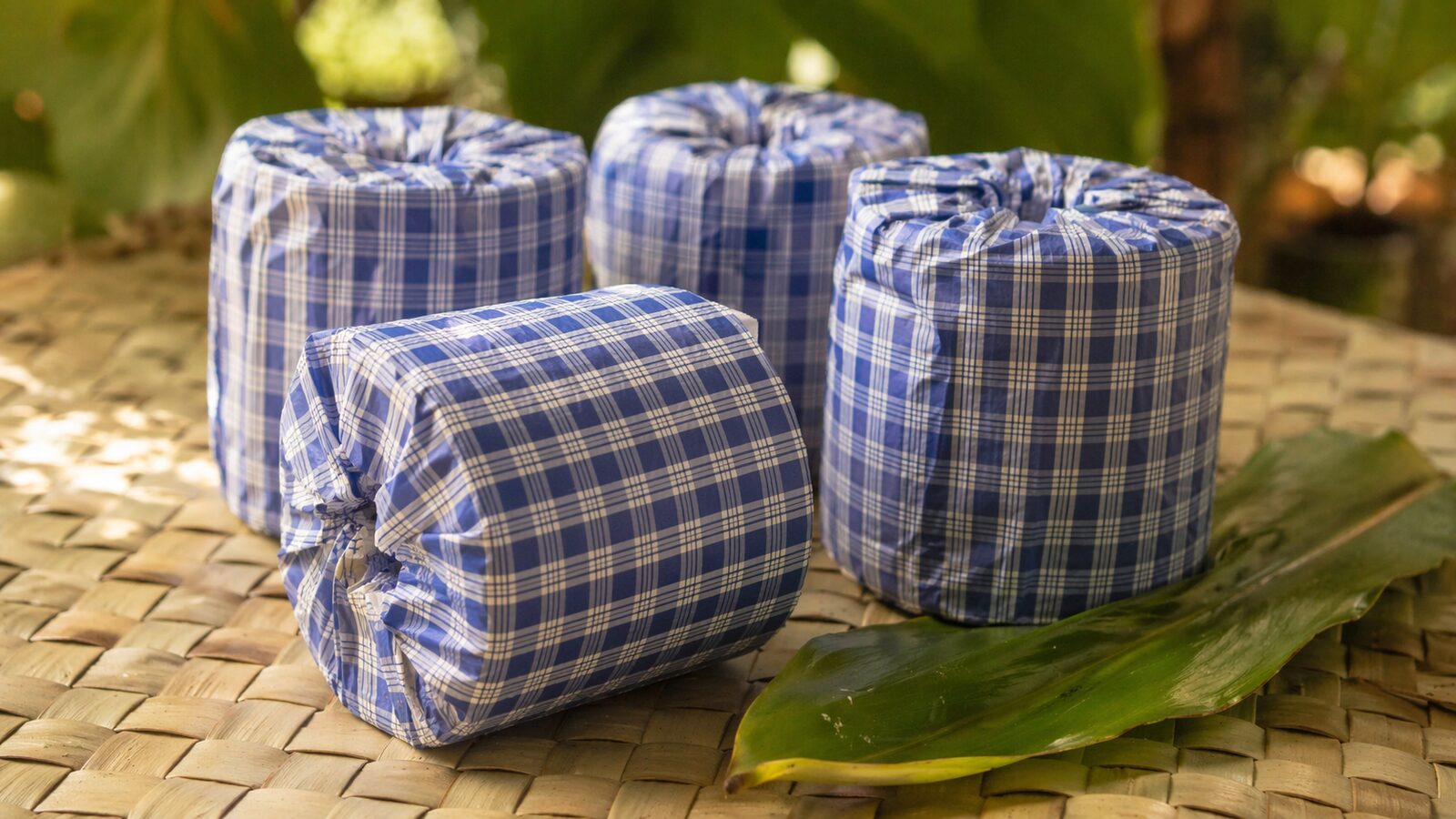 Four palaka-wrapped toilet paper rolls on a woven lauhala mat with a ti leaf